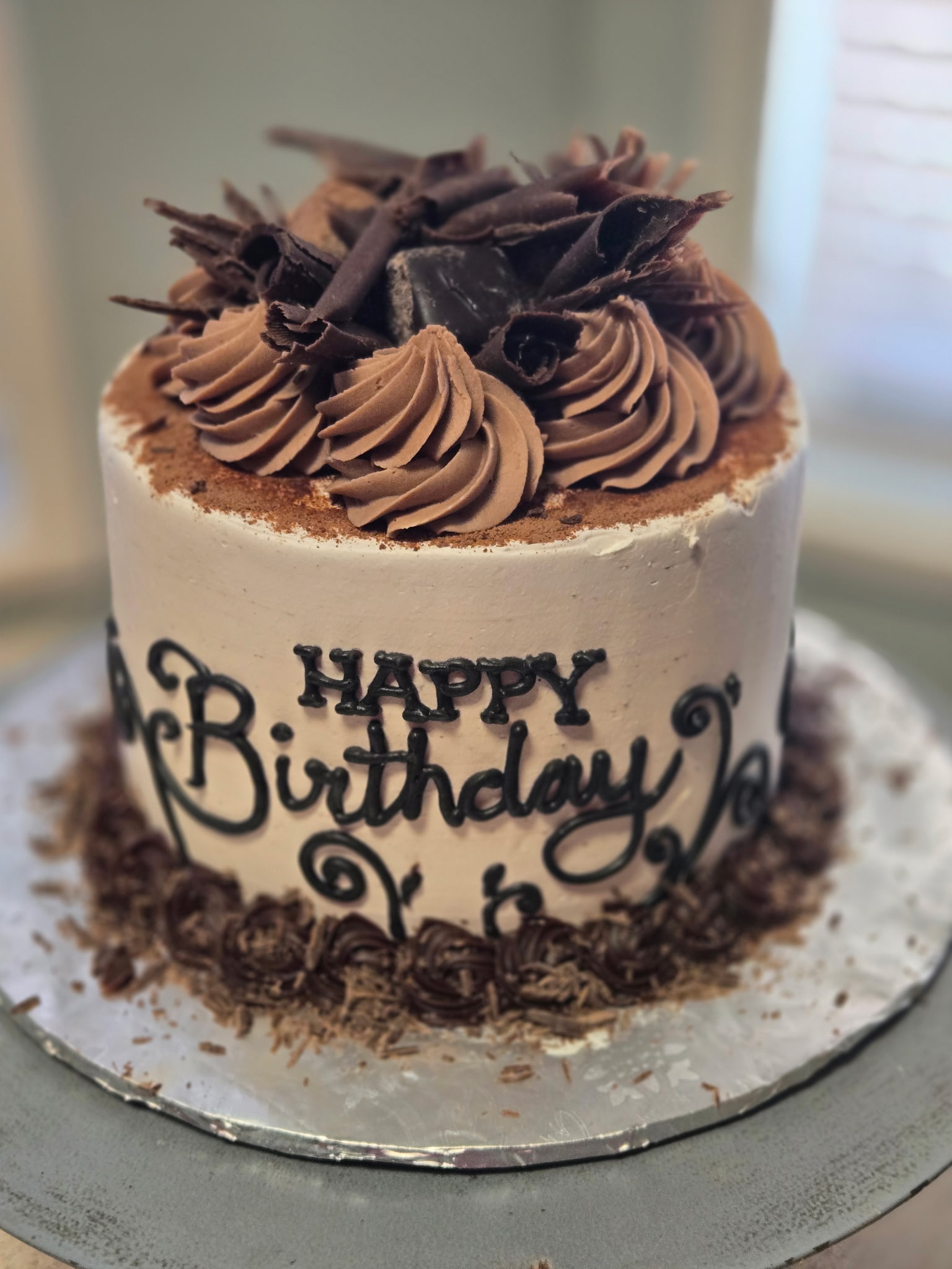 Single-tier birthday cake with white buttercream, dark-chocolate "Happy Birthday" piped in cursive, and a topper of chocolate buttercream rosettes and chocolate curls.