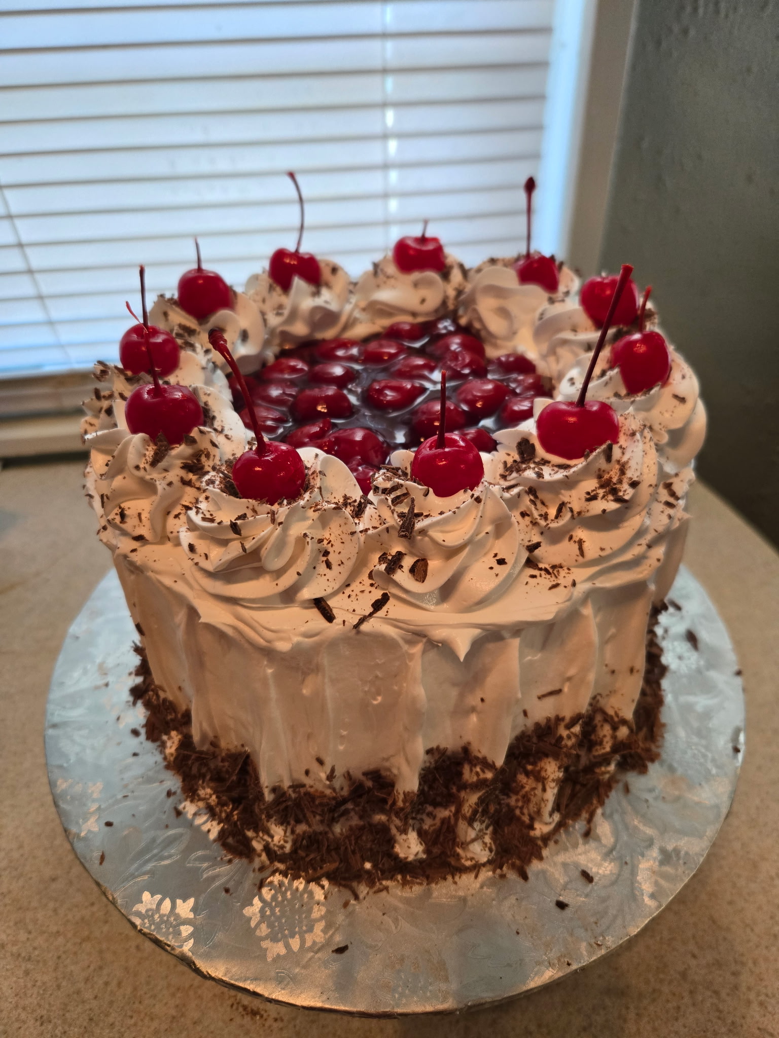Black Forest cake with whipped-cream rosettes, fresh cherries on stems, a pool of cherry filling on top, and a chocolate-shaving border at the base.