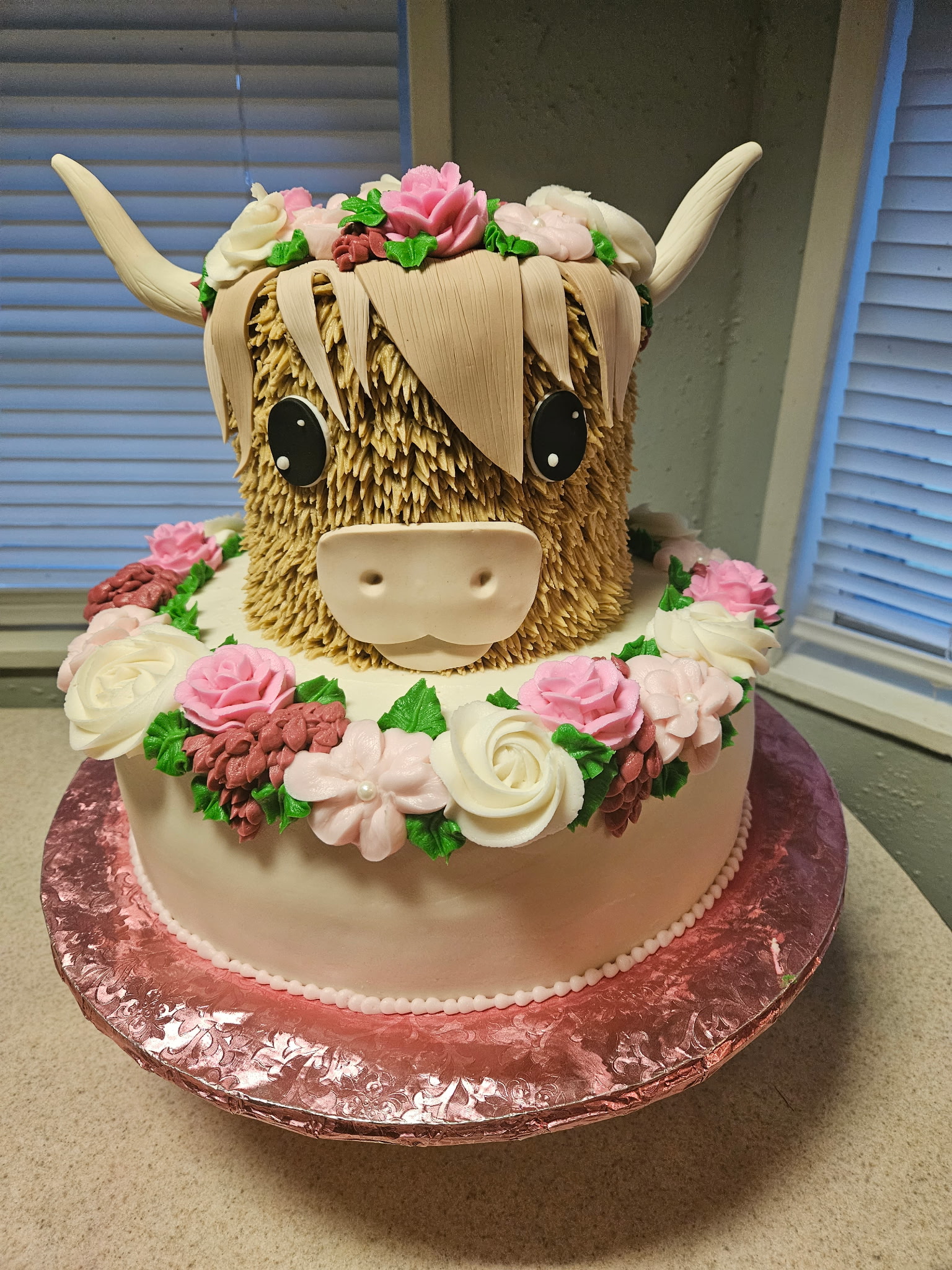 Two-tier Highland cow themed birthday cake with a sculpted fondant cow's head topper and a wreath of hand-piped buttercream roses around the base.