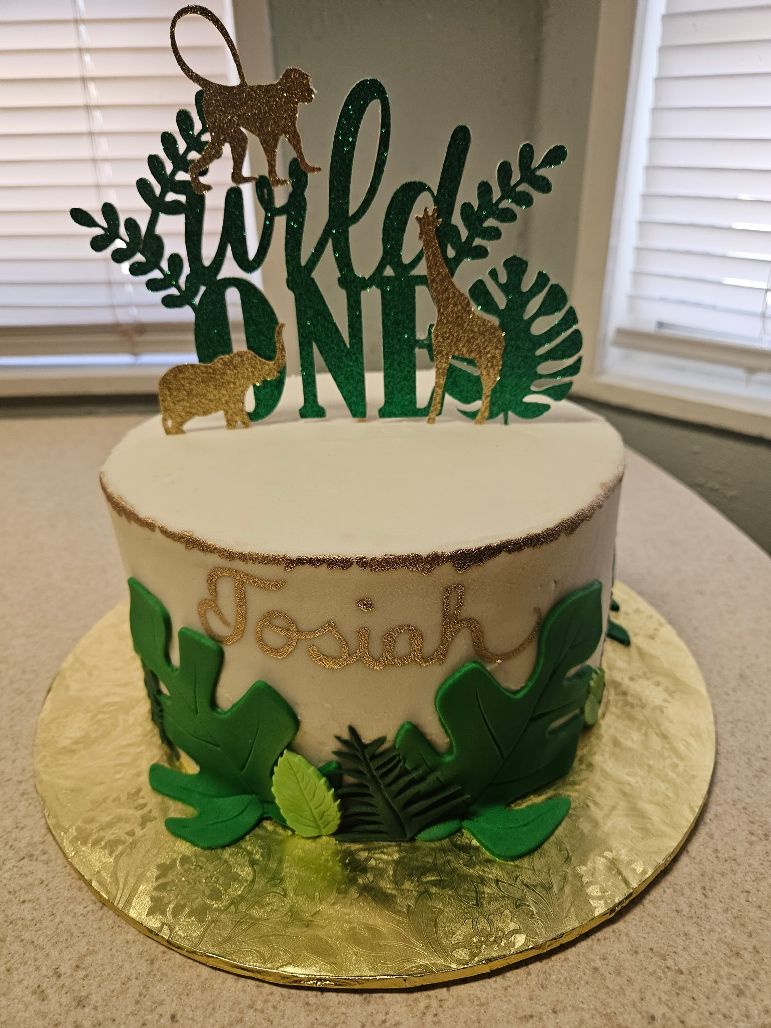 First-birthday "Wild ONE" jungle cake with cream buttercream, gold piped name, green fondant leaves at the base, and a safari-animal silhouette topper.