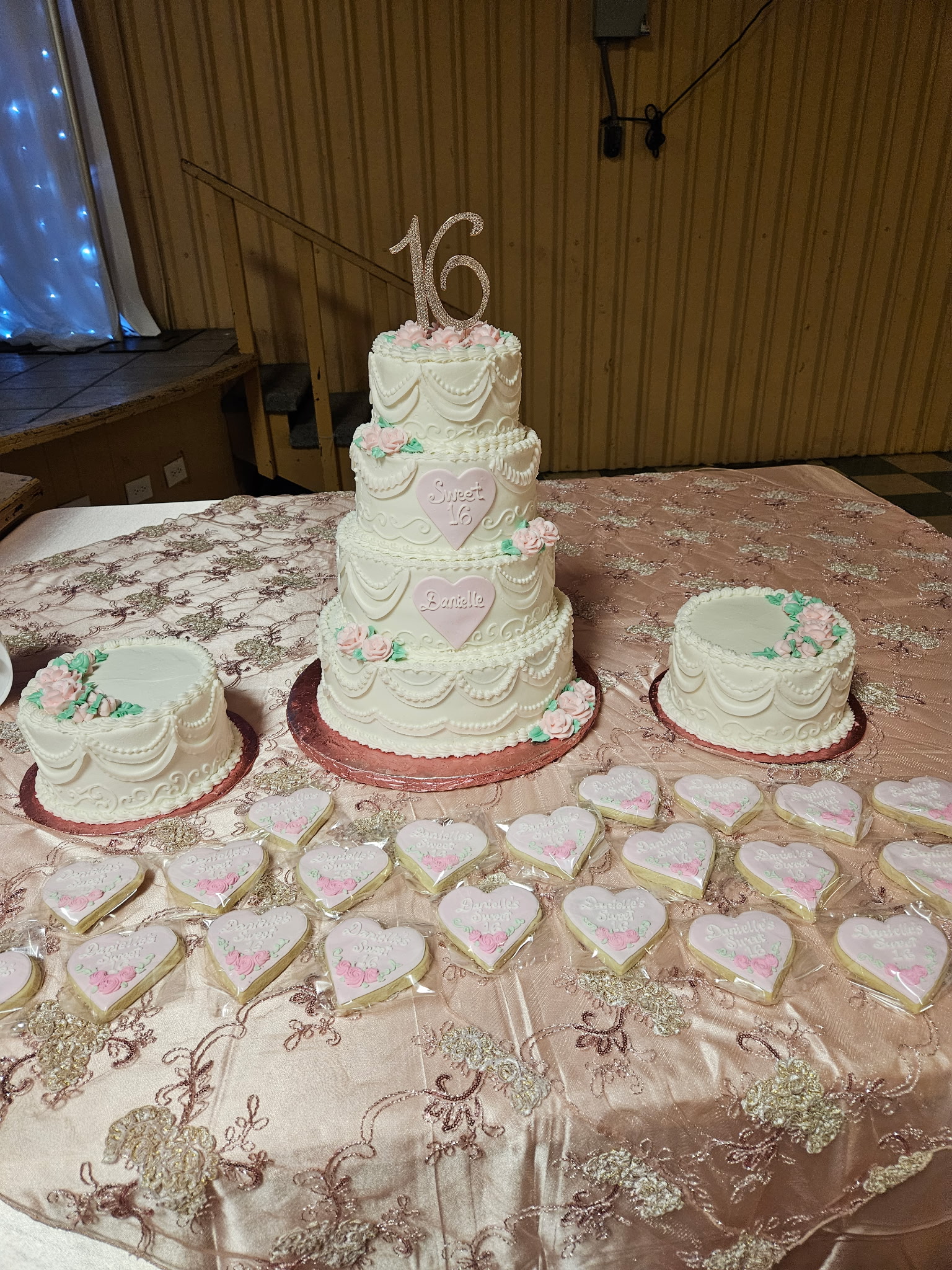 Tiered Quinceañera cake in white buttercream with draped scrollwork, pink heart medallions on each tier, and a tray of matching pink heart sugar cookies.
