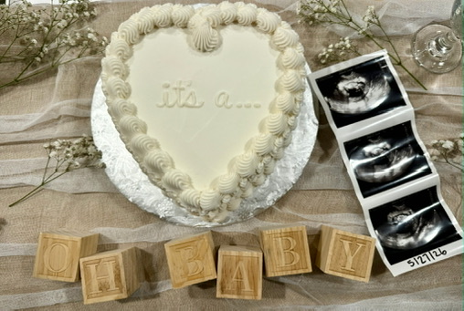 Heart-shaped gender-reveal cake in cream buttercream with "it's a…" piped in the center, styled beside an ultrasound photo strip.
