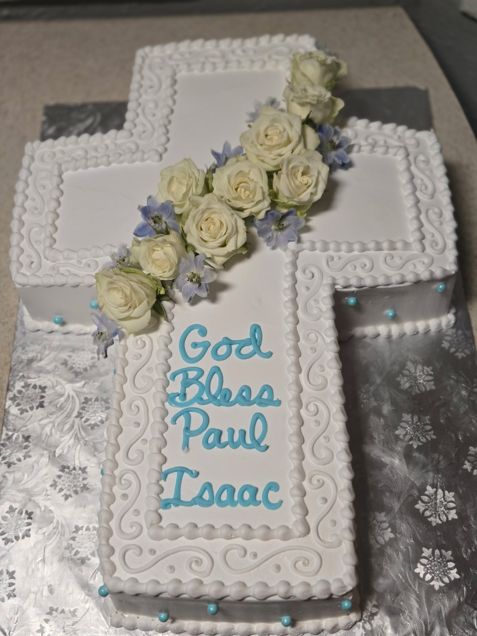 Cross-shaped First Communion cake in white buttercream with hand-piped scrollwork borders, a diagonal spray of fresh white roses, and blue piped lettering.