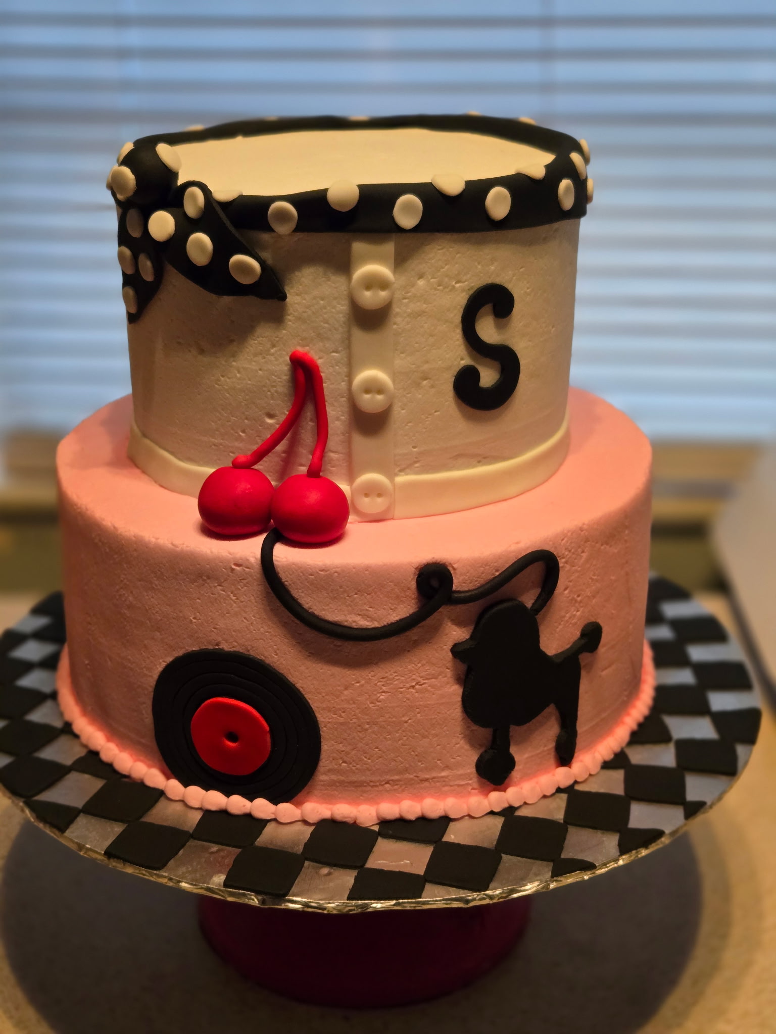 Two-tier 1950s sock-hop birthday cake with a black-and-white poodle-skirt top tier and a pink bottom tier featuring a piped poodle, vinyl record, and checkered base.