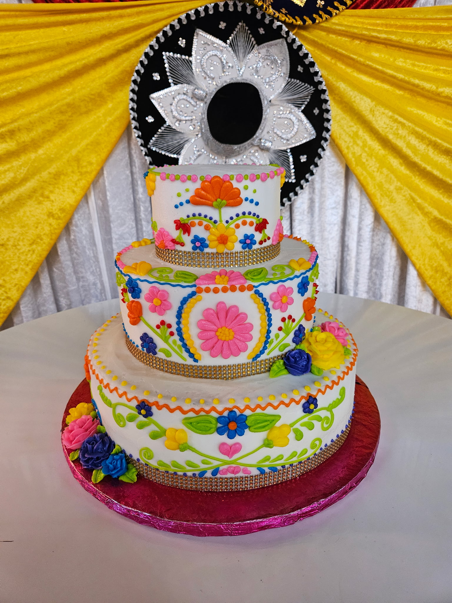 Three-tier Quinceañera cake hand-piped with bright Mexican floral embroidery and a decorative sombrero topper.